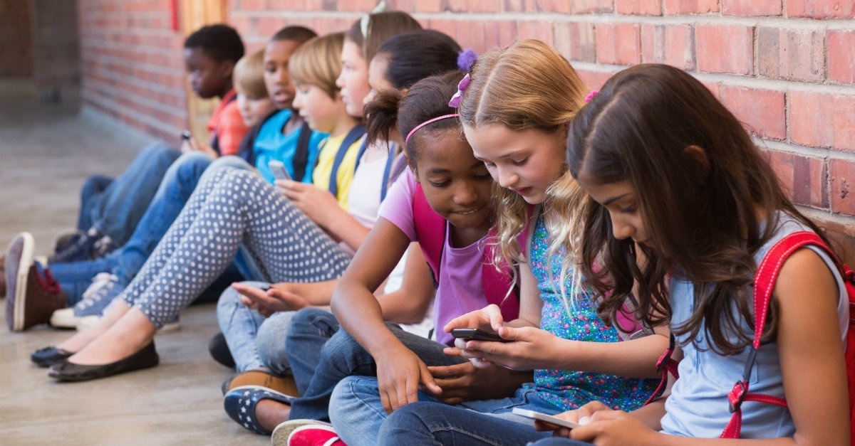 A group of kids all sitting against the wall on their phones