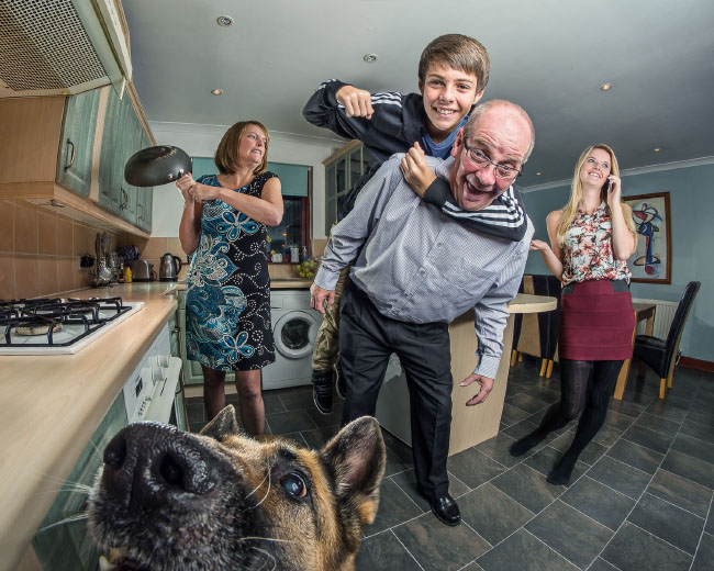 A son on top of his dad, a dog in the bottom, a lady with a pan ready to swing, and another lady on her phone