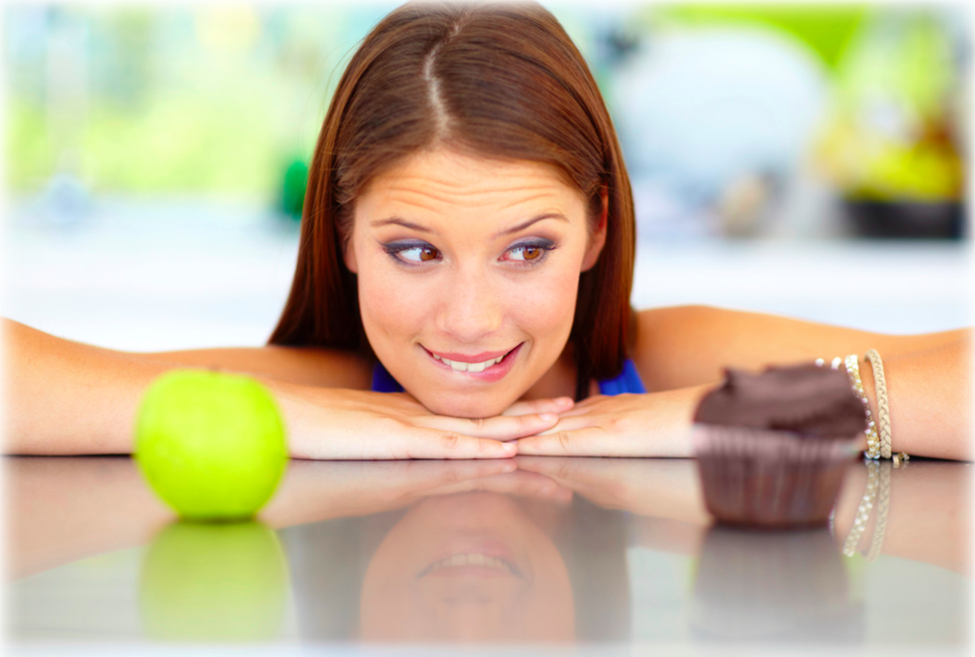 A lady leaning on a table debating between eating a green apple or a chocolate cup cake
