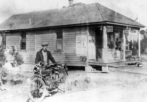 A black and white photo of a man standing with his bike in front of an ice cream parlor