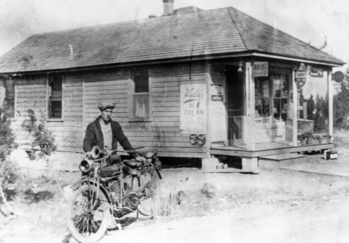 A black and white photo of a man standing with his bike in front of an ice cream parlor