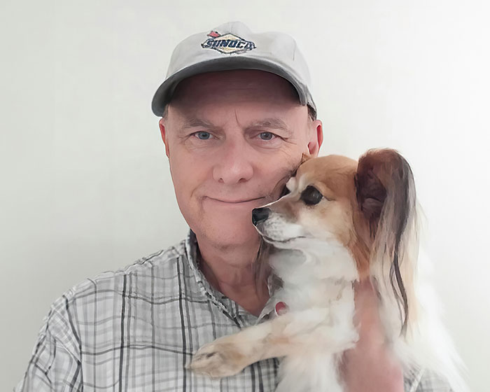 An older man in a sunoco hat, and a button down shirt, holding his little dog.