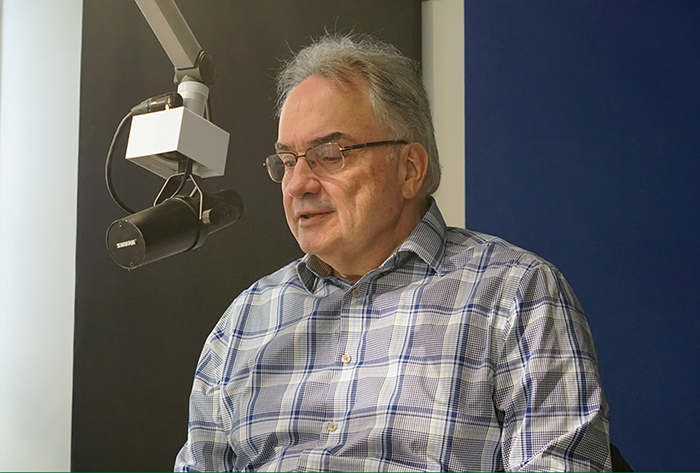 An older gentleman sitting in front of a mic, wearing a button down shirt and glasses
