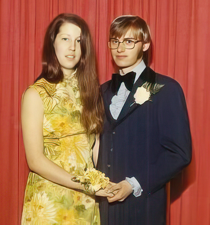 A couple standing together in front of a red curtain, the man is wearing a suit and the woman is wearing a yellow dress