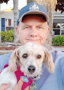 A man in a hat and button-down shirt with his dog, in a pink harness