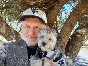 A man in a hat and jacket, holding his dog, who is in a sweater