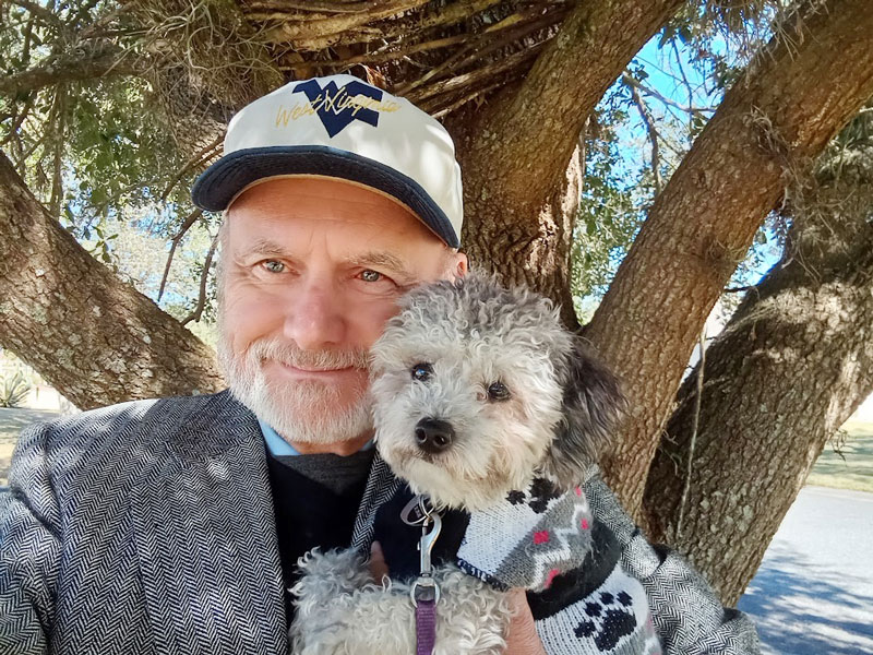 A man in a hat and jacket, holding his dog, who is in a sweater