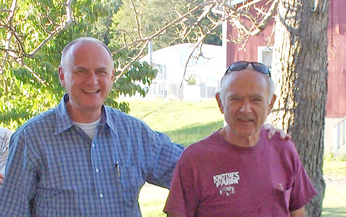 Two men stand outdoors, smiling while one wears sunglasses. A green landscape and a red barn are visible in the background.
