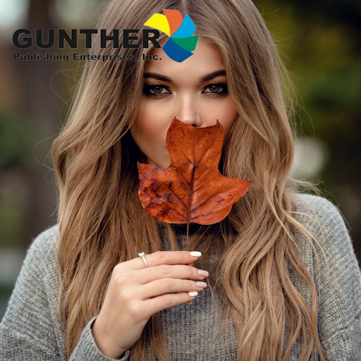 A person holds an autumn leaf in front of their face, showcasing long, wavy hair and wearing a cozy gray sweater in a natural setting.
