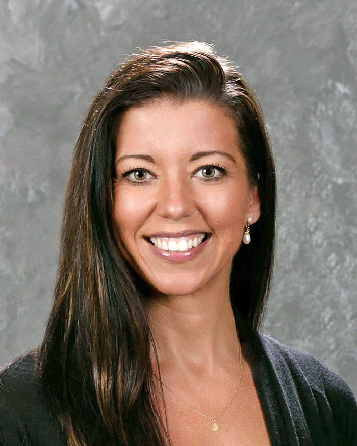 A woman with long, wavy hair wearing a black cardigan and a delicate necklace against a textured gray background.